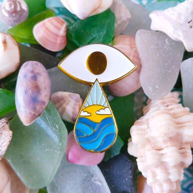 an enamel pin depicting a stylized eye with a teardrop that contains an ocean horizon.