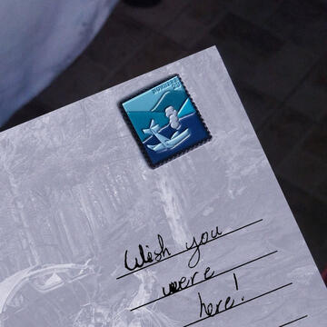 an enamel pin depicting a stamp of a plane crash on a postcard. the postcard reads wish you were here